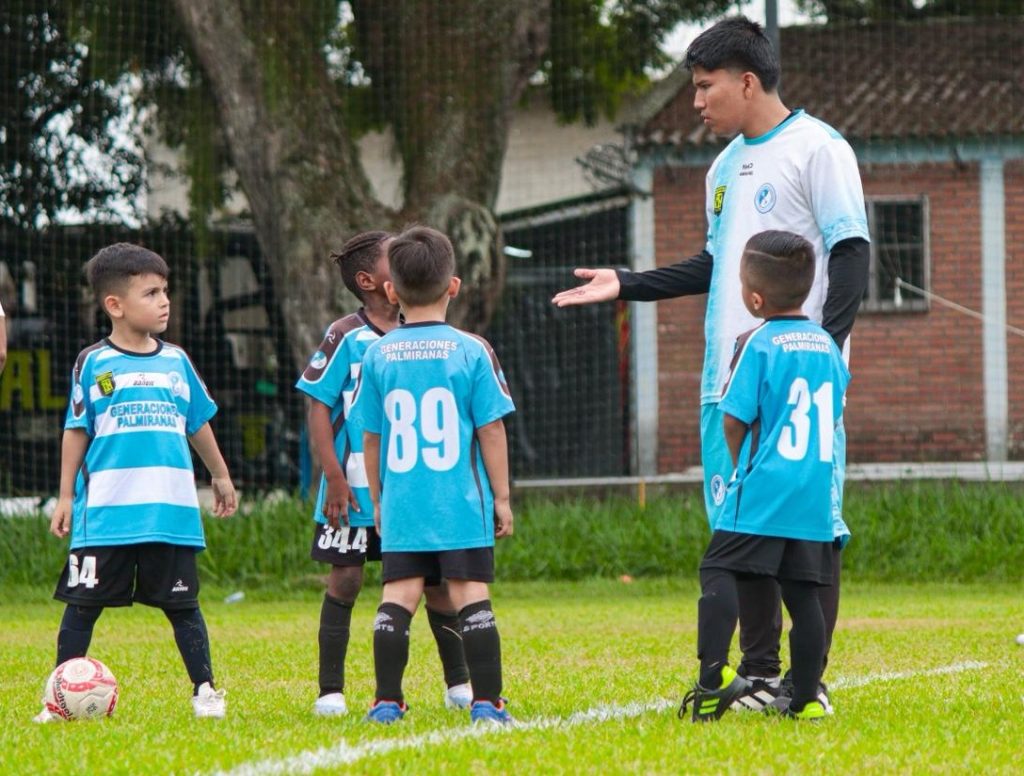 Fotografía en plano medio corto en una cancha de césped. Un entrenador joven, de pie a la derecha, gesticula con la mano mientras se dirige a un grupo de cuatro niños pertenecientes a un equipo de fútbol infantil. Los pequeños deportistas, de espaldas y de perfil, visten uniformes de franjas azules y blancas con la inscripción "Generaciones Palmiranas" y escuchan atentamente las indicaciones.