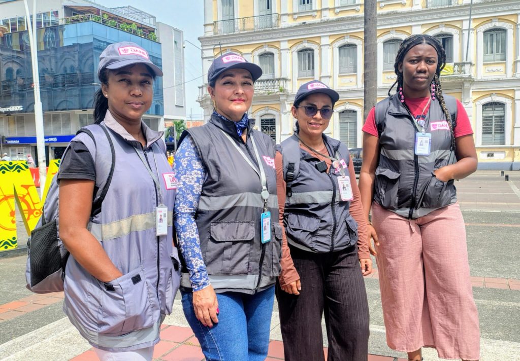 Fotografía de cuatro mujeres encuestadoras del DANE en una plaza pública, vistiendo chalecos grises, gorras de la institución y escarapelas de identificación