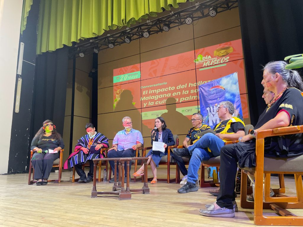 Panel de discusión en un escenario teatral con siete personas sentadas en semicírculo, debatiendo frente a una gran pantalla que anuncia un conversatorio sobre la cultura Malagana por el Día Internacional de los Museos.