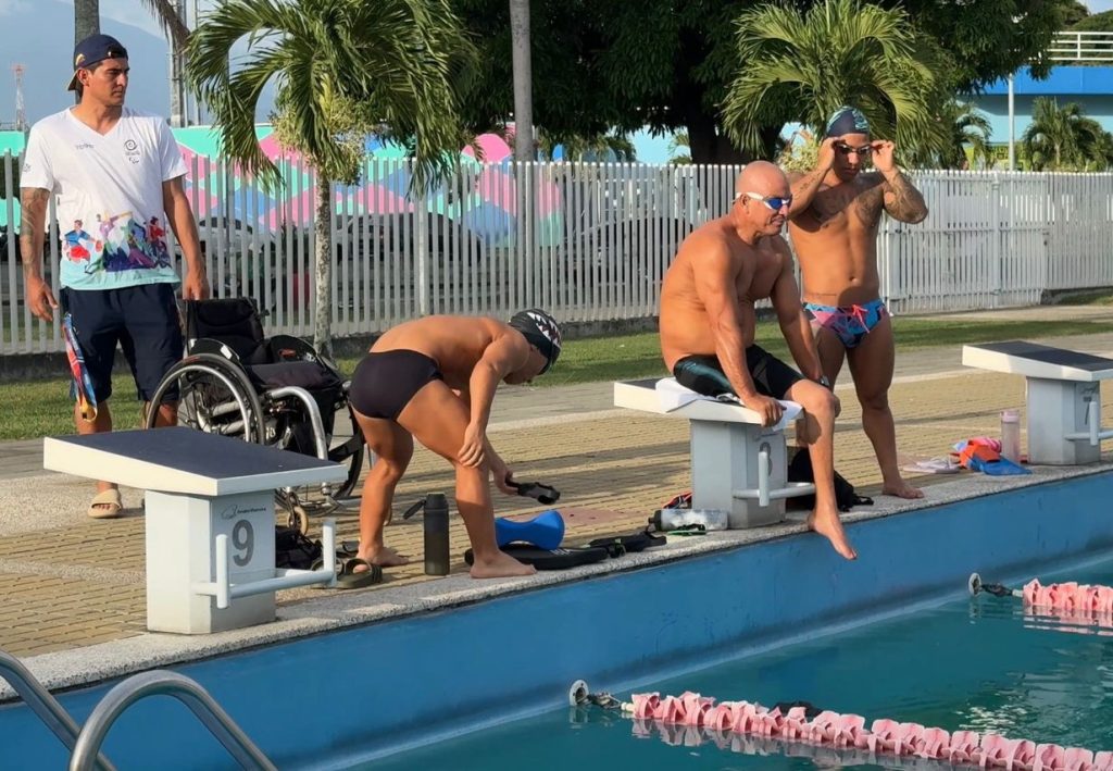 Fotografía de deportistas de natación adaptada en el borde de una piscina olímpica exterior, preparándose junto a un entrenador y una silla de ruedas.