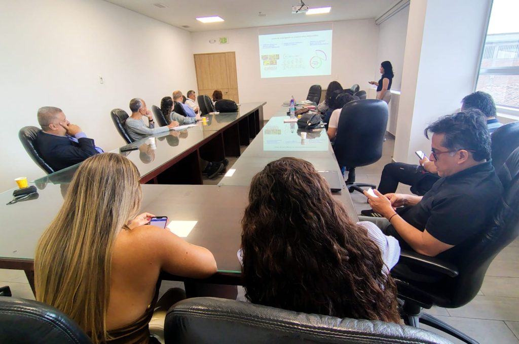 Reunión de trabajo en una sala de juntas de la Alcaldía de Palmira. Un grupo de personas se encuentra sentado alrededor de una gran mesa modular en forma de U. Al fondo, una mujer de pie realiza una presentación apoyada en la proyección de una diapositiva en la pared. En primer plano, varias personas atienden la sesión y revisan sus dispositivos móviles.