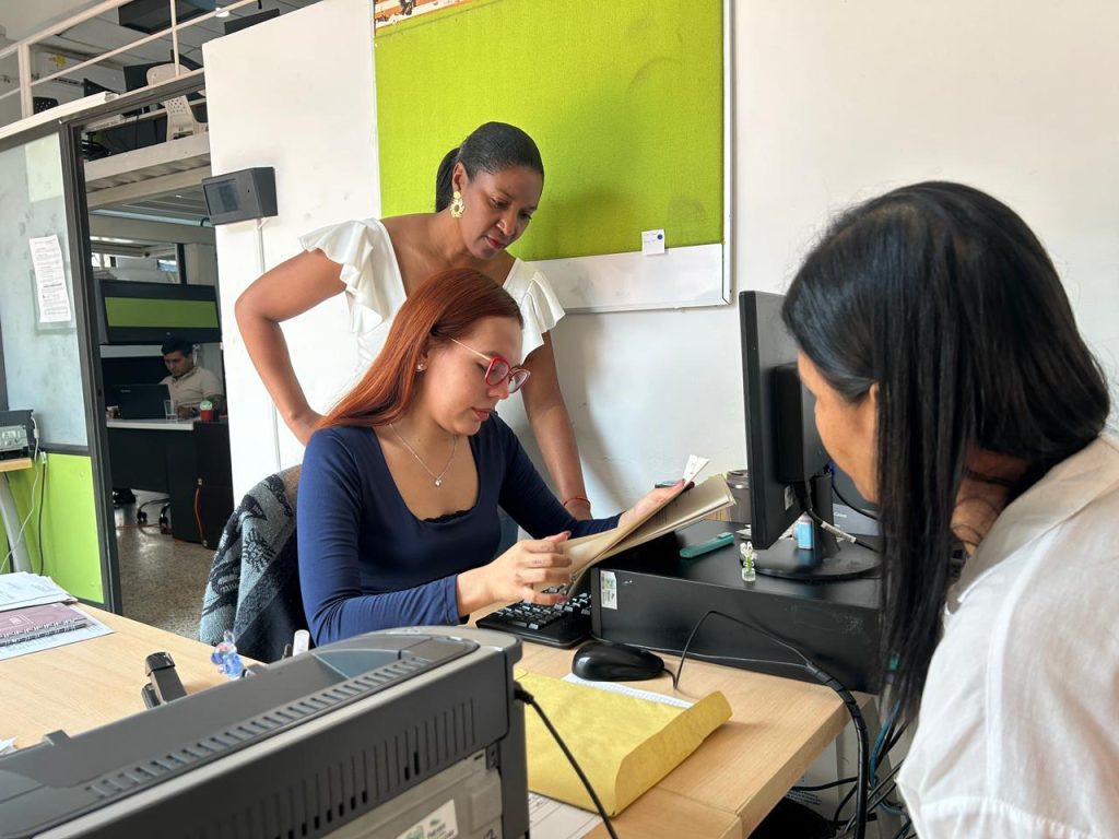 Tres personas trabajan en una oficina de la Alcaldía de Palmira. En primer plano, una persona sentada de espaldas revisa un monitor. En el centro, una mujer con gafas y cabello cobrizo analiza unos documentos impresos junto al teclado. Detrás de ella, otra mujer afrodescendiente con blusa blanca observa detalladamente los papeles sobre el escritorio. Al fondo se aprecia el entorno laboral con archivadores y otra estación de trabajo.