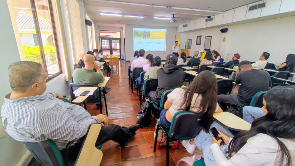 Fotografía en plano general de un salón de clases universitario donde un grupo diverso de personas asiste a una presentación. Al fondo, un expositor habla de pie junto a una pantalla de proyección con diapositivas. El público está sentado en filas de pupitres individuales orientados hacia el frente. En primer plano, se observa de perfil a varias personas prestando atención y utilizando sus dispositivos móviles.