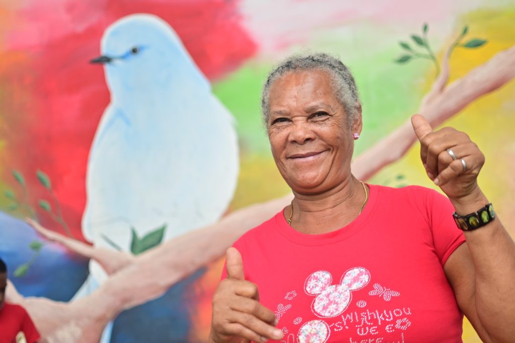 Fotografía en plano medio de una persona mayor sonriente, con cabello corto y canoso, que viste una camiseta roja. Tiene ambos pulgares hacia arriba frente a un mural colorido que muestra un ave azul sobre una rama.