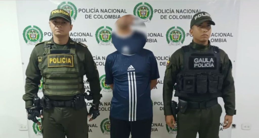 Fotografía de tres personas de pie frente a un fondo institucional de la Policía Nacional de Colombia. En los extremos se encuentran dos integrantes de la fuerza policial con uniformes verdes, chalecos tácticos y gorras. En el centro, una persona civil con el rostro difuminado viste una camiseta deportiva azul marino con líneas blancas.