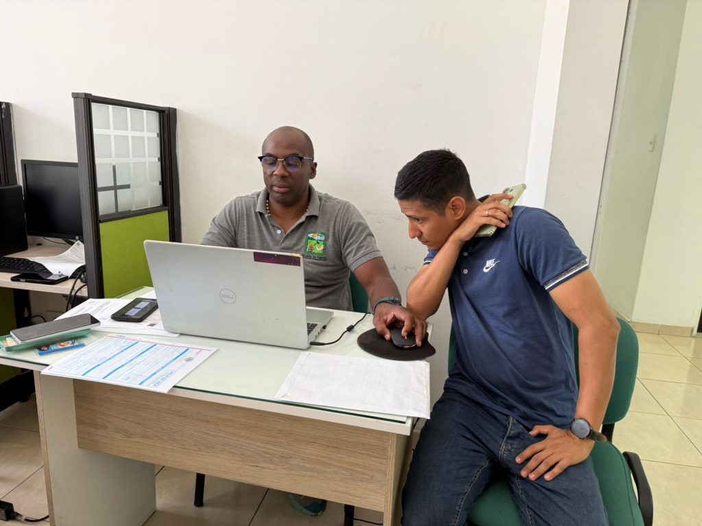 Dos personas están sentadas en un escritorio en una oficina de la Alcaldía de Palmira. A la izquierda, un servidor público con gafas y polo gris con el logo institucional mira hacia un computador portátil Dell abierto. A la derecha, otra persona con polo azul y jeans se inclina para mirar la pantalla mientras habla por un teléfono móvil que sostiene con su mano derecha. Sobre el escritorio hay documentos, una calculadora y un ratón. El fondo muestra una partición de oficina.