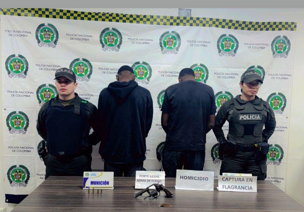Dos integrantes de la Policía Nacional de Colombia custodian a dos personas detenidas, quienes se encuentran de espaldas frente a un telón institucional con logotipos de la policía. En primer plano, sobre una mesa, se exhiben un arma de fuego, cartuchos y carteles informativos que indican los motivos de las capturas.