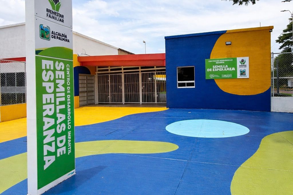 Fachada del Centro de Desarrollo Infantil Semillas de Esperanza en Palmira. Incluye un tótem informativo verde y blanco a la izquierda y un edificio al fondo con paredes pintadas de amarillo y azul. El suelo exterior es un patio de juegos multicolor con formas pintadas en tonos azules, verdes y amarillos