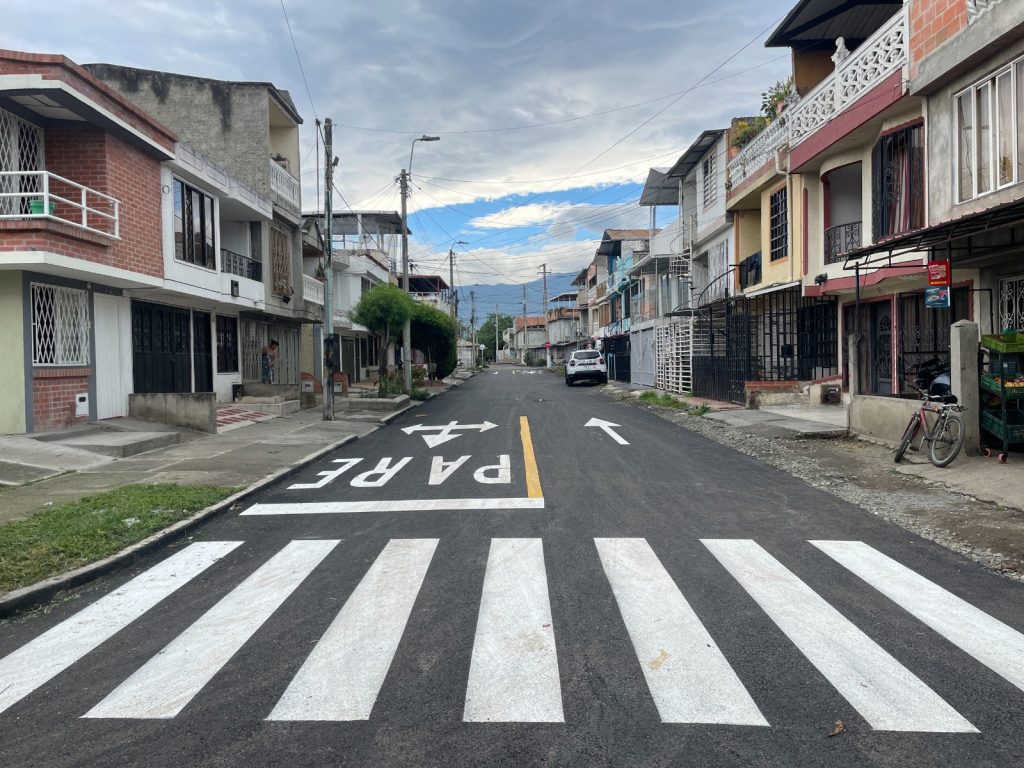Calle residencial asfaltada en perspectiva con un paso peatonal de líneas blancas en primer plano y la palabra "PARE" pintada en el suelo junto a flechas de dirección. A los lados hay casas de dos y tres pisos, postes de luz, vegetación y un automóvil blanco estacionado al fondo