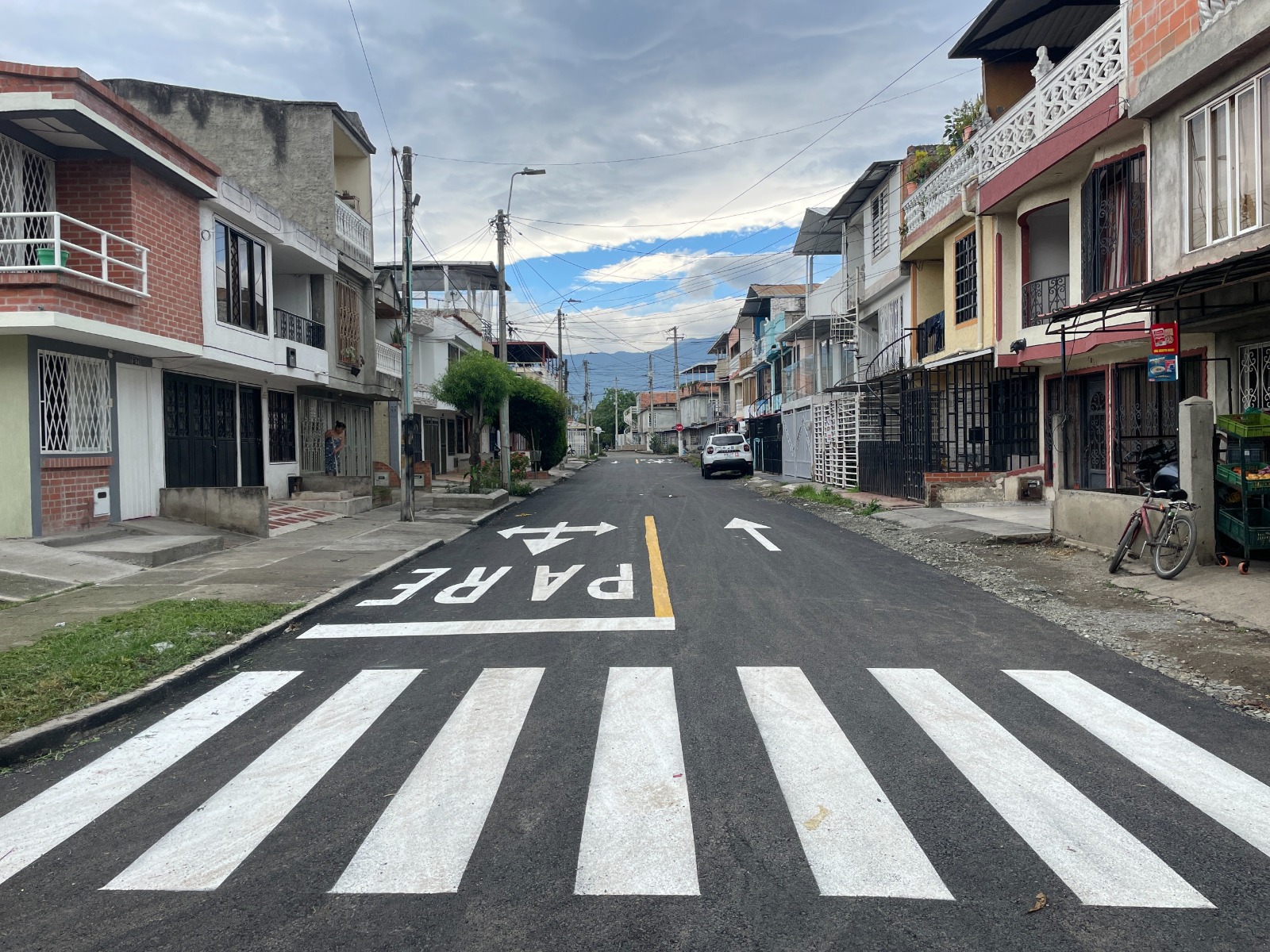 Calle residencial asfaltada en perspectiva con un paso peatonal de líneas blancas en primer plano y la palabra "PARE" pintada en el suelo junto a flechas de dirección. A los lados hay casas de dos y tres pisos, postes de luz, vegetación y un automóvil blanco estacionado al fondo