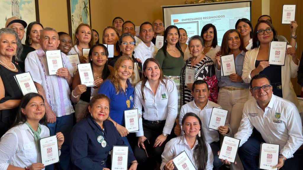 Fotografía grupal de personas adultas sonriendo en un salón interior mientras sostienen diplomas o cartillas blancas con un logotipo circular rojo. Al fondo se observa una pantalla de proyección con el título "EMPRESARIOS RECONOCIDOS" y un documento institucional.