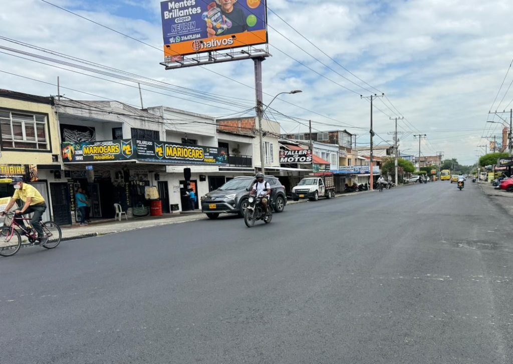 Calle urbana pavimentada con comercios locales a la izquierda, personas transitando en bicicleta y motocicleta, y un gran cartel publicitario elevado al fondo bajo un cielo parcialmente nublado.