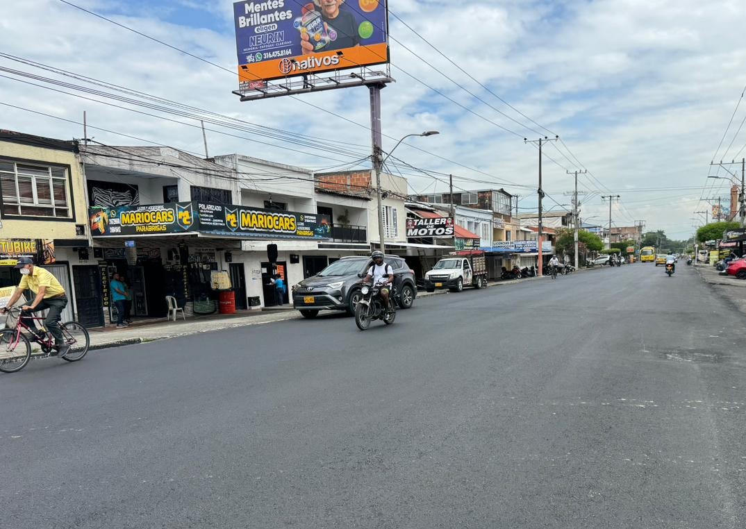 Calle urbana pavimentada con comercios locales a la izquierda, personas transitando en bicicleta y motocicleta, y un gran cartel publicitario elevado al fondo bajo un cielo parcialmente nublado.