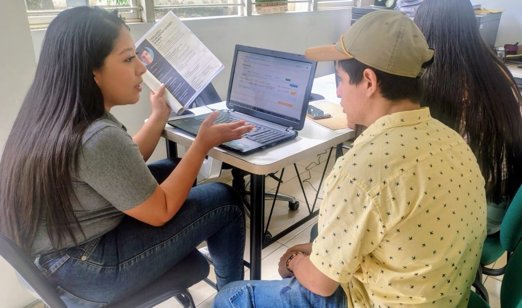 Una funcionaria con camiseta gris asesora a un joven con gorra beige frente a un computador portátil en una oficina. La mujer sostiene una hoja de vida impresa con una fotografía mientras gesticula explicando el contenido de la pantalla. Ambos están sentados a los lados de un escritorio blanco. Al fondo se observa a otra persona de espaldas.
