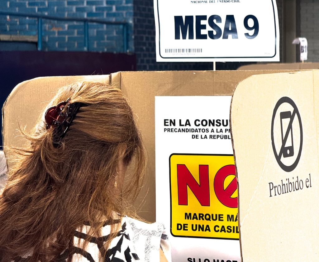 Primer plano desde atrás de una persona con cabello castaño y largo recogido con una pinza, mientras ejerce su derecho al voto dentro de un cubículo de votación de cartón. Arriba del cubículo se lee un cartel que dice "MESA 9" de la Registraduría Nacional del Estado Civil. En el panel lateral hay un símbolo que indica la prohibición del uso de teléfonos móviles.