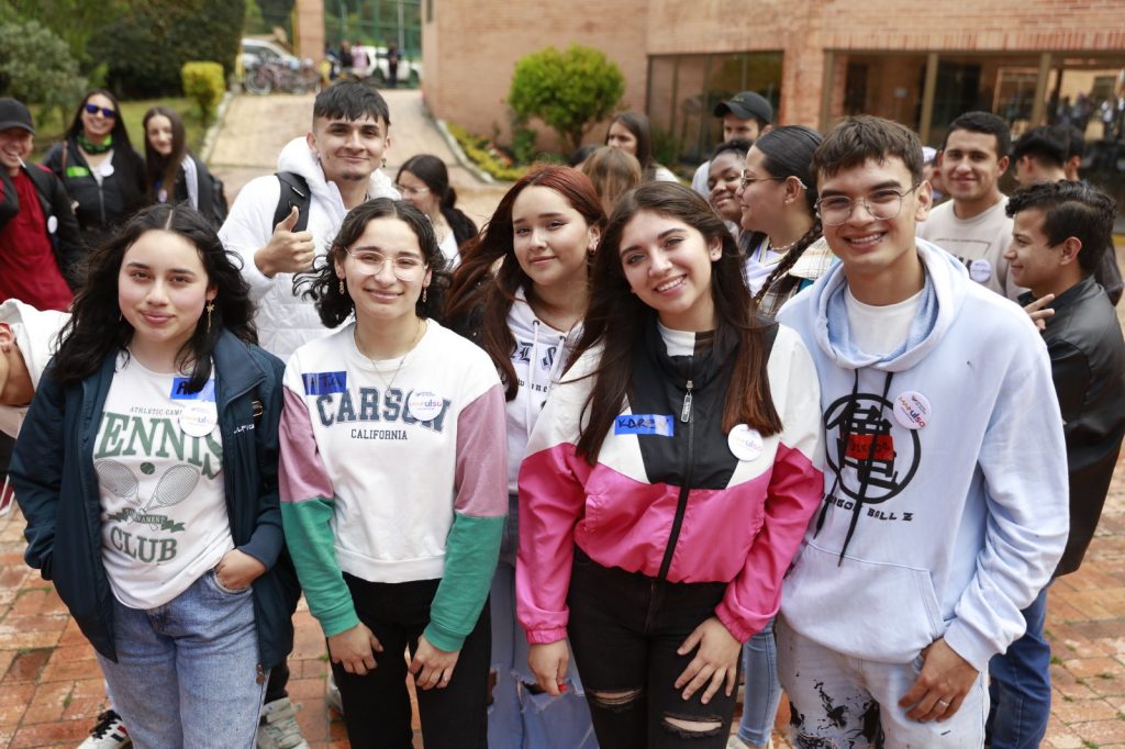 Un grupo numeroso de personas jóvenes posa sonriente en un espacio abierto con suelo de adoquines frente a un edificio de ladrillo. En primer plano se destacan cuatro integrantes vistiendo ropa casual, buzos y chaquetas deportivas; algunas llevan escarapelas adhesivas de un evento institucional. Al fondo, más participantes acompañan la actividad en una atmósfera comunitaria.