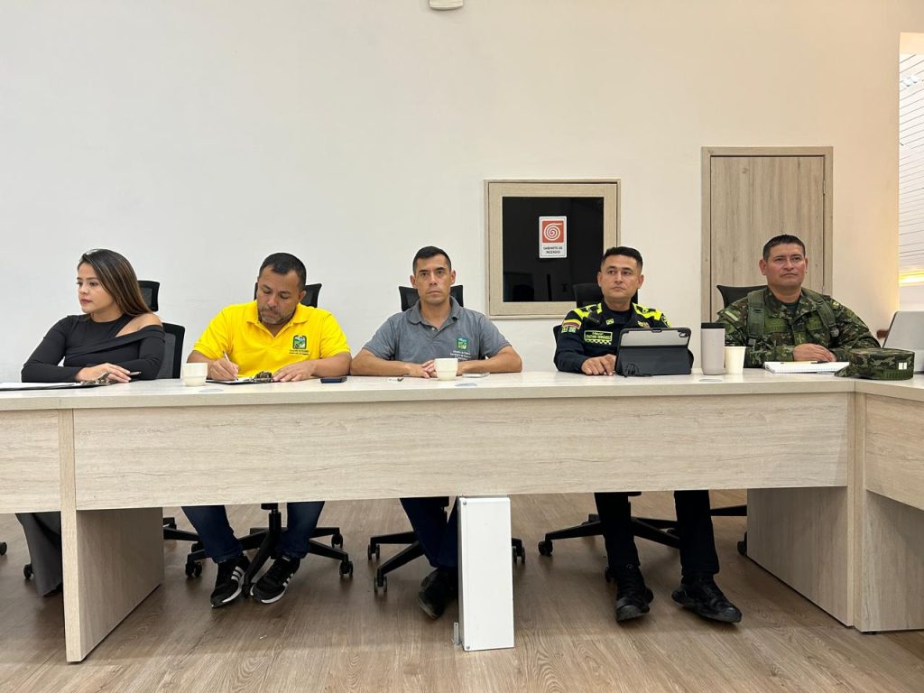 Cinco personas están sentadas en línea detrás de una mesa de juntas clara durante una reunión de seguimiento. De izquierda a derecha: una mujer con prenda negra, un hombre con polo amarillo institucional tomando notas, un hombre con polo gris de la Alcaldía, un integrante de la Policía Nacional con una tableta y un integrante del Ejército Nacional con uniforme camuflado. Al fondo se aprecia una pared blanca con un gabinete de incendios y una puerta de madera.