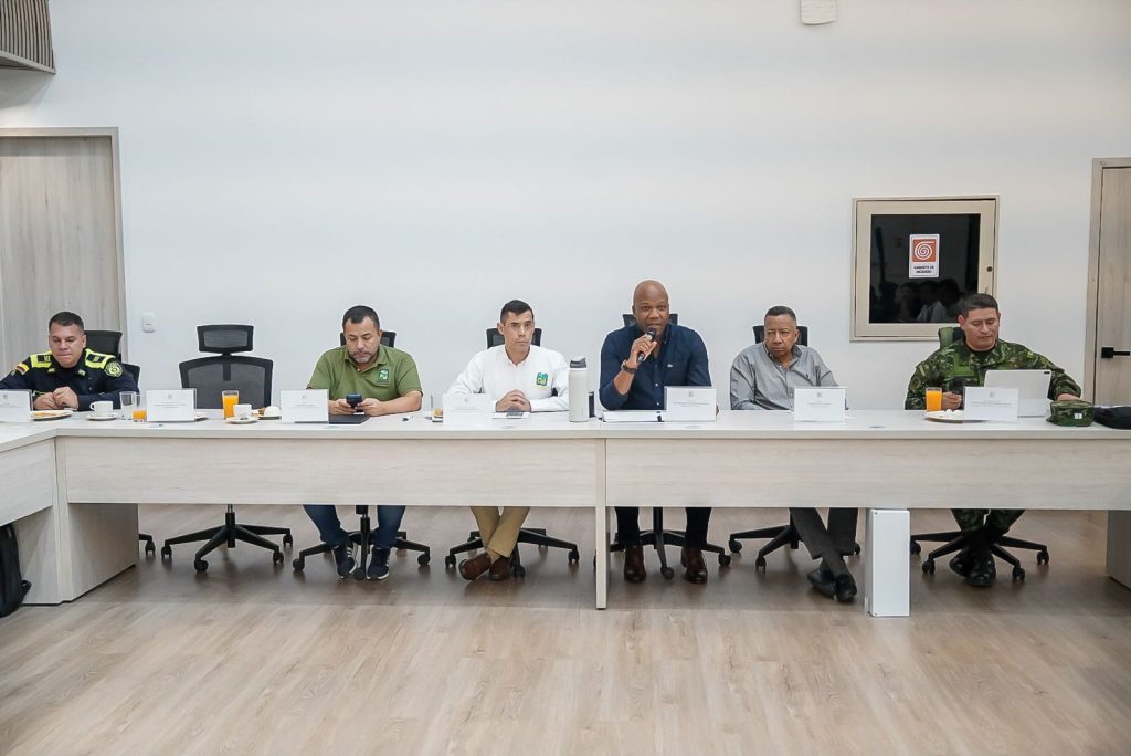 Siete personas están sentadas en línea detrás de una mesa de juntas clara durante una reunión institucional. En el centro, el alcalde Víctor Ramos habla sosteniendo un micrófono. A sus lados se encuentran funcionarios de la Alcaldía, delegados civiles, un integrante de la Policía Nacional en el extremo izquierdo y un integrante del Ejército Nacional con uniforme camuflado en el extremo derecho. Sobre la mesa hay identificadores institucionales, bebidas y computadores portátiles.