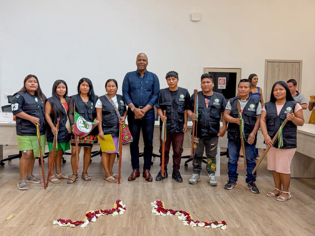 Fotografía grupal del alcalde de Palmira, Víctor Ramos, junto a integrantes de Resguardo Indígena, quienes visten chalecos institucionales negros y portan bastones de mando ceremoniales. En el suelo se aprecia un arreglo con pétalos de flores.
