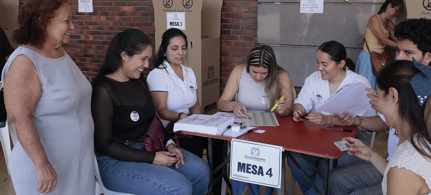 Fotografía de un grupo de personas en una mesa de votación de la Registraduría Nacional del Estado Civil. Cuatro personas sentadas atienden la mesa número 4, revisando documentos y planillas con actitud sonriente. Dos personas están sentadas al frente esperando, mientras una mujer de pie a la izquierda observa la escena con una sonrisa. Al fondo se aprecian cubículos de votación de cartón.