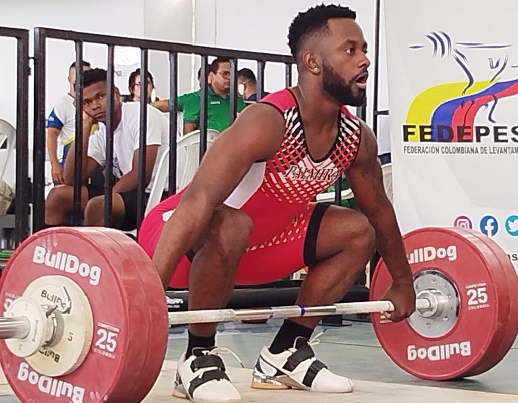 Un pesista afrodescendiente con uniforme rojo de Palmira en posición de cuclillas sostiene una barra con grandes discos rojos marca BullDog durante una competencia de levantamiento de pesas.