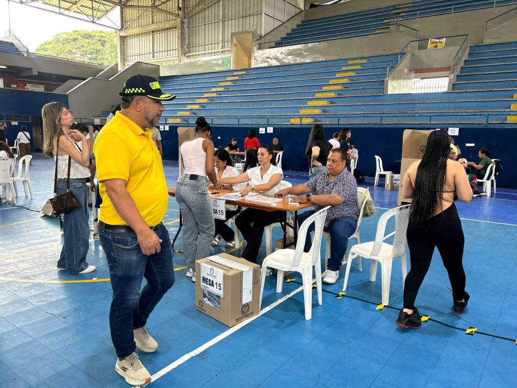 Vista del interior de un coliseo deportivo acondicionado como puesto de votación, con jurados atendiendo una mesa electoral y el secretario de gobierno de la alcaldía caminando en primer plano.