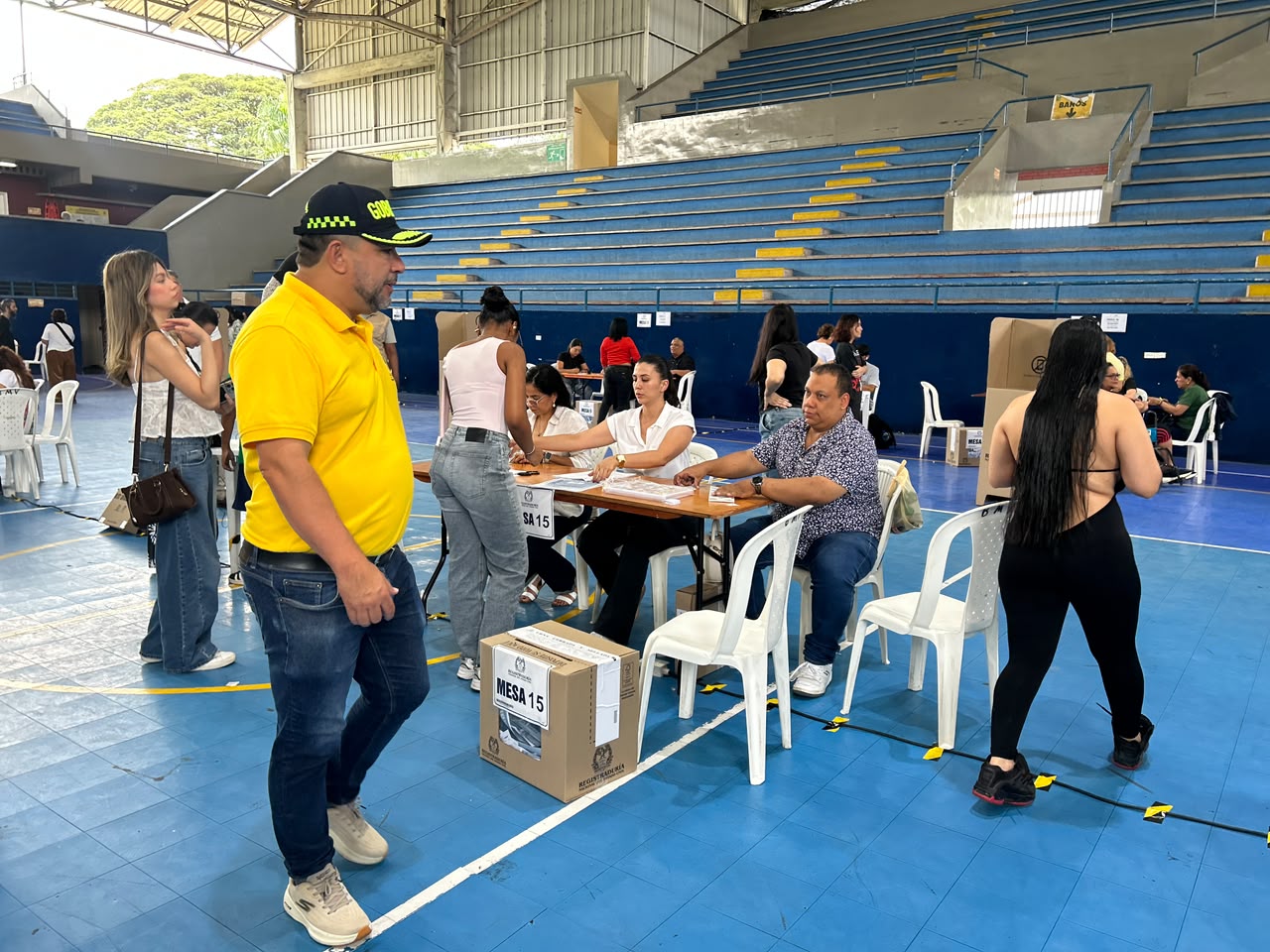 Vista del interior de un coliseo deportivo acondicionado como puesto de votación, con jurados atendiendo una mesa electoral y el secretario de gobierno de la alcaldía caminando en primer plano.