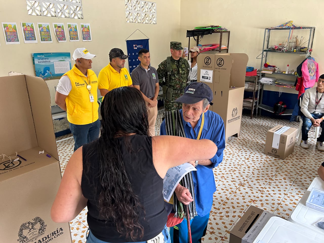 Un hombre de la tercera edad con camisa azul y gorra recibe asistencia de una mujer para votar en una mesa de votación, rodeado de jurados, observadores electorales y un militar en un aula escolar.