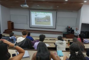 Un profesor de pie frente a una clase universitaria durante una presentación. La proyección muestra a un grupo de personas en un entorno rural con el título 'El Magueyal, estado de Hidalgo, México'. Estudiantes de diversas identidades observan desde sus bancos