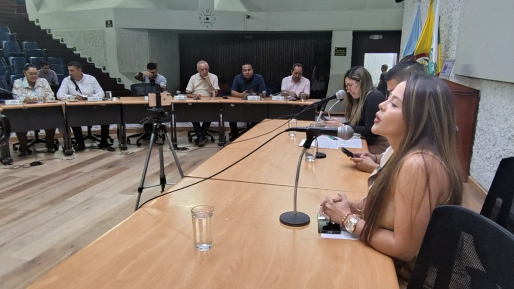 En primer plano, a la derecha, Secretaria General de la Alcaldía de Palmira habla frente a un micrófono sobre una mesa de madera clara. Detrás de ella hay otra mujer y un hombre. Al fondo, varios concejales asistentes están sentados en una mesa semicircular escuchando la intervención