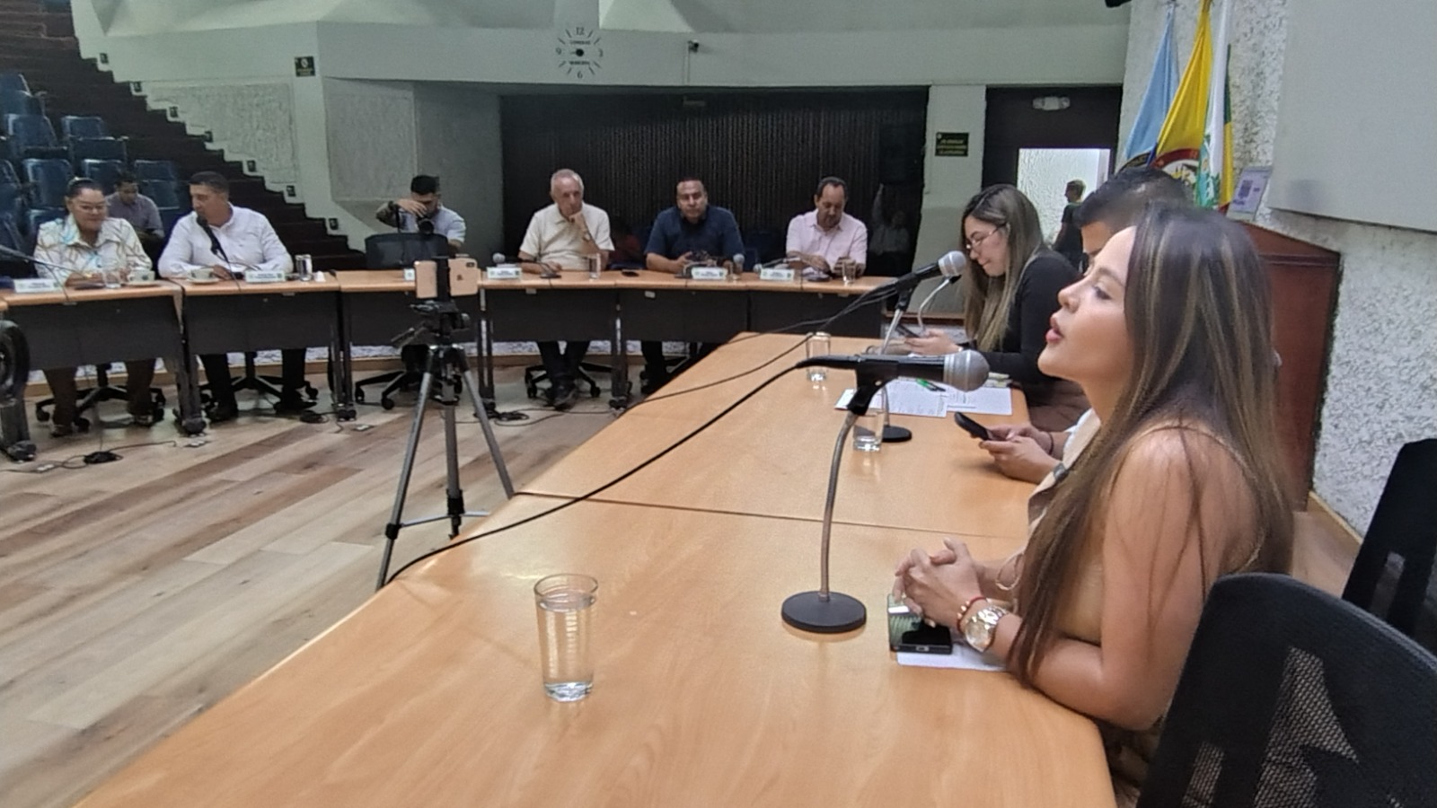 En primer plano, a la derecha, Secretaria General de la Alcaldía de Palmira habla frente a un micrófono sobre una mesa de madera clara. Detrás de ella hay otra mujer y un hombre. Al fondo, varios concejales asistentes están sentados en una mesa semicircular escuchando la intervención