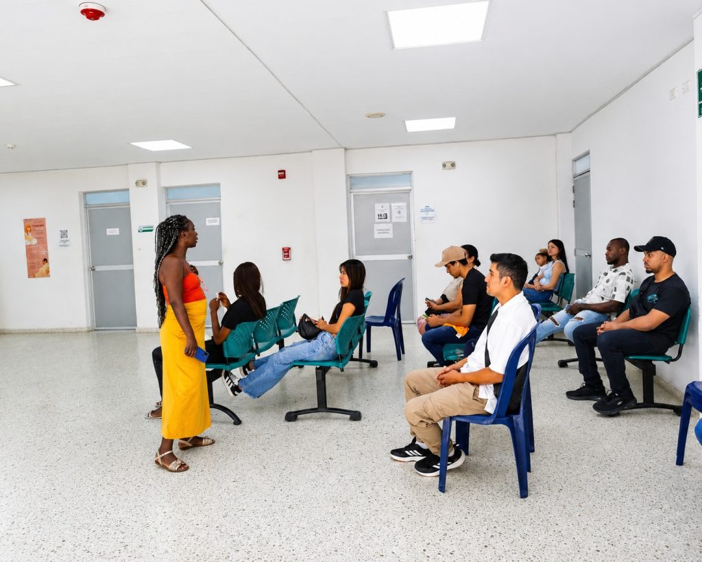 Grupo de personas sentadas en una sala de espera médica mientras una mujer de pie con falda amarilla conversa con una de las usuarias en un entorno clínico iluminado.