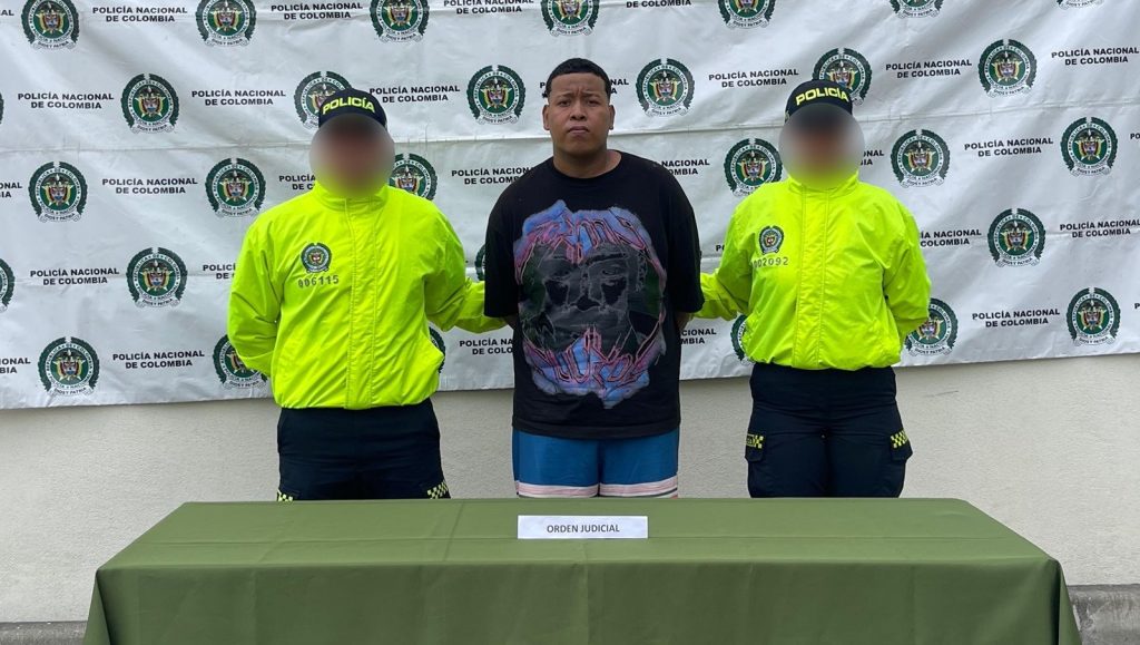 Persona bajo custodia policial, flanqueada por dos agentes de la Policía Nacional de Colombia con chaquetas verdes de alta visibilidad, delante de un fondo institucional con el logo de la entidad