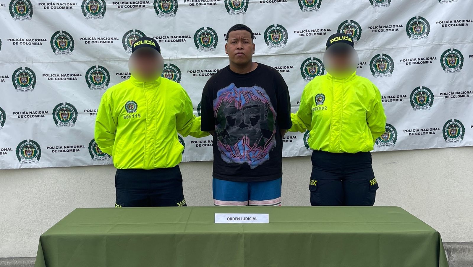 Persona bajo custodia policial, flanqueada por dos agentes de la Policía Nacional de Colombia con chaquetas verdes de alta visibilidad, delante de un fondo institucional con el logo de la entidad