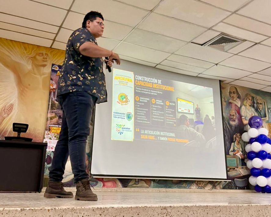 Una persona de perfil da una presentación en un escenario frente a una gran pantalla de proyección. Viste una camisa estampada de manga corta, pantalones oscuros y botas. La diapositiva proyectada habla sobre la "Construcción de Capacidad Institucional" de la Alcaldía de Palmira