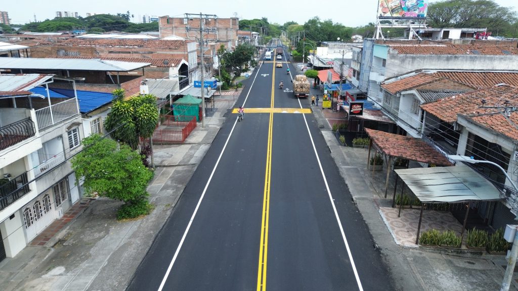 Vista aérea de una calle urbana residencial recién pavimentada y señalizada. El asfalto oscuro muestra líneas dobles amarillas en el centro, líneas blancas en los costados y un resalto peatonal señalizado en amarillo. A los lados hay viviendas de dos pisos, comercios locales, árboles y algunos vehículos en movimiento, como motocicletas y un camión al fondo.