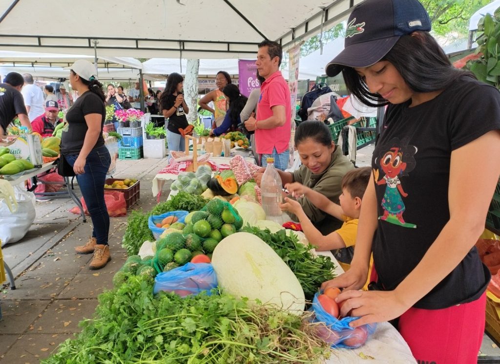 Personas de la comunidad compran y venden hortalizas, verduras y frutas frescas bajo carpas blancas en una feria campesino local.