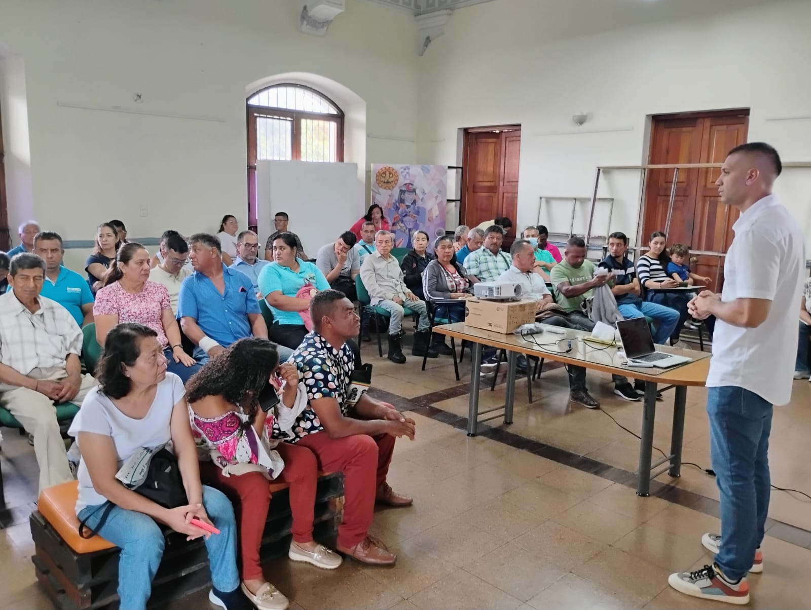 Un grupo diverso de personas asiste a una reunión comunitaria en un salón amplio. Al frente, un hombre con camisa blanca y jeans expone de pie ante la audiencia.