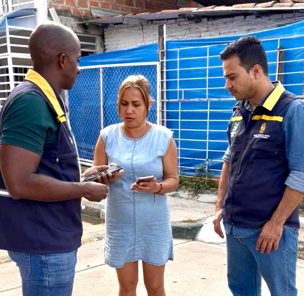 Dos personas con chalecos institucionales azules y amarillos conversan en la calle con una mujer que sostiene un teléfono móvil.