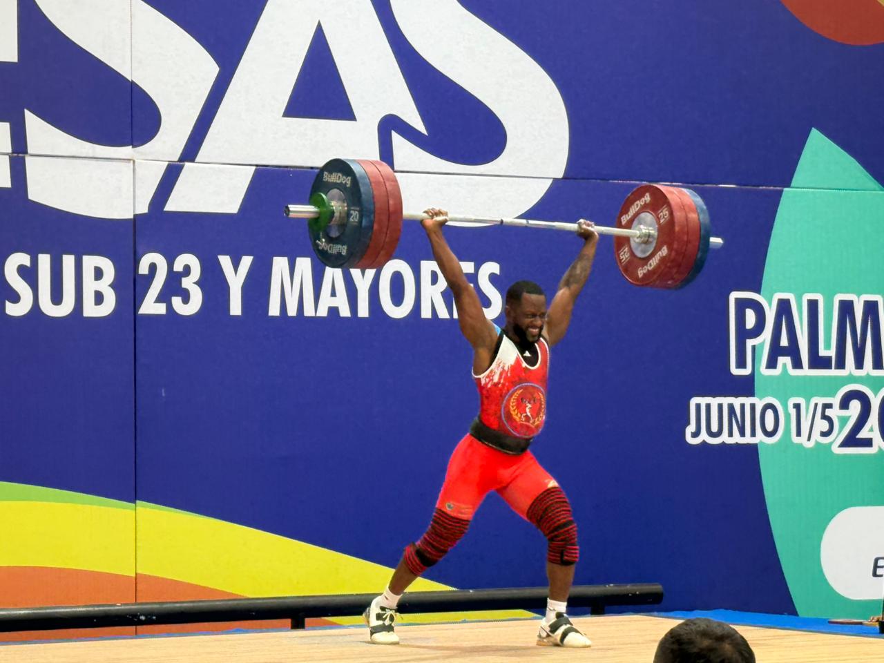 Una persona deportista de levantamiento de pesas sostiene una barra cargada con discos sobre su cabeza durante una competencia oficial. El fondo es de color azul con letras blancas.