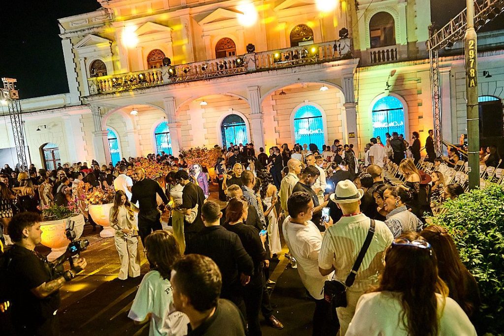 Fotografía de un evento nocturno de moda al aire libre con una gran concurrencia de personas socializando frente al edificio Antigua Estación del Ferrocarril de Palmira iluminada con luces amarillas, turquesas y violetas.