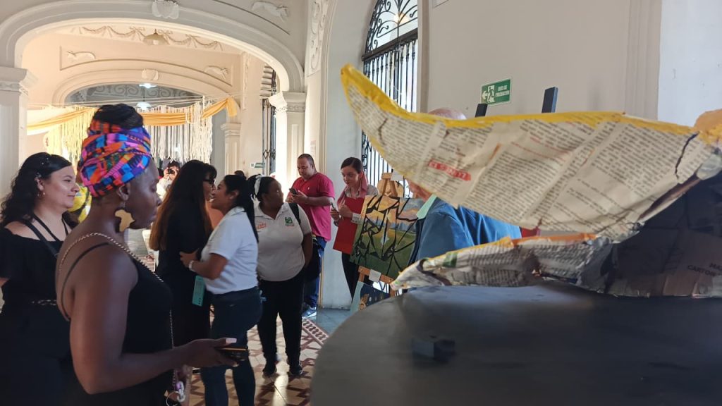 En imagen personas asistentes recorren una exposición de arte de la EMA en un pasillo histórico de la Alcaldía de Palmira, observando pinturas y esculturas en papel maché