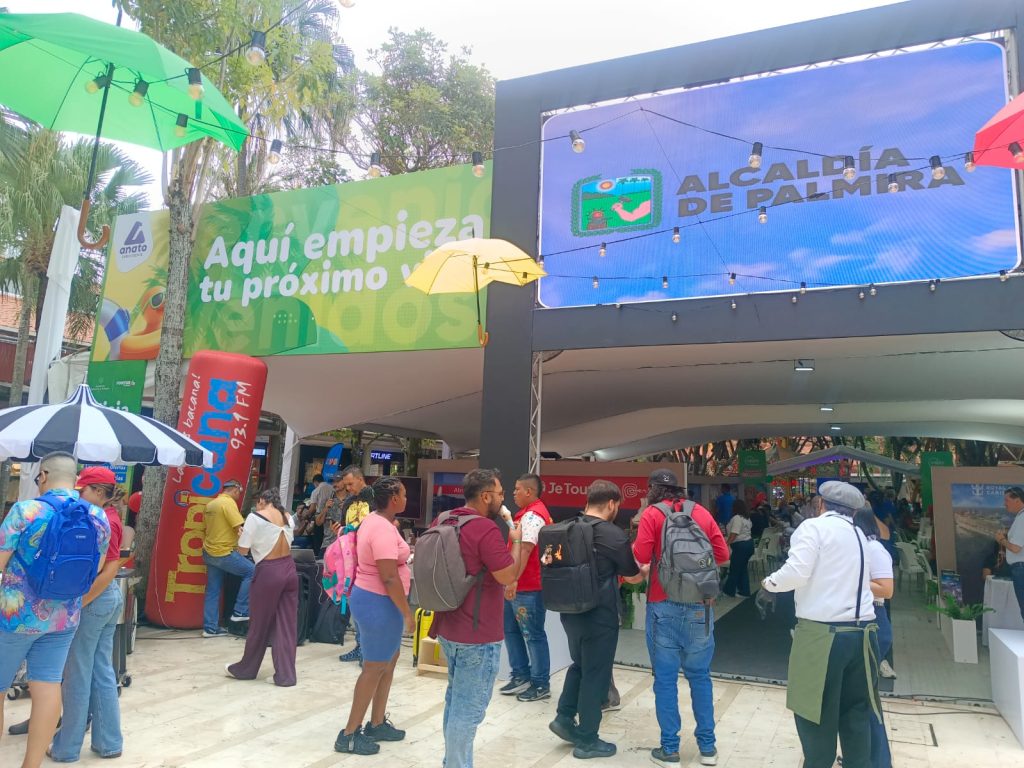 Diversas personas caminan y conversan frente a un gran arco de entrada. Un cartel verde arriba dice "Aquí empieza tu próximo viaje". A la derecha, una pantalla digital muestra el logo de la "Alcaldía de Palmira". Hay sombrillas de colores, vegetación de fondo y puestos promocionales en un ambiente concurrido y diurno.