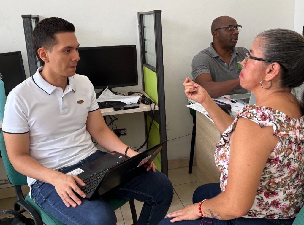 Una mujer de mediana edad se comunica mediante Lengua de Señas con un funcionario de la Alcaldía de Palmira, quien la atiende con un computador portátil en su regazo dentro de una oficina de atención ciudadana. Al fondo, otro funcionario atiende en su escritorio.