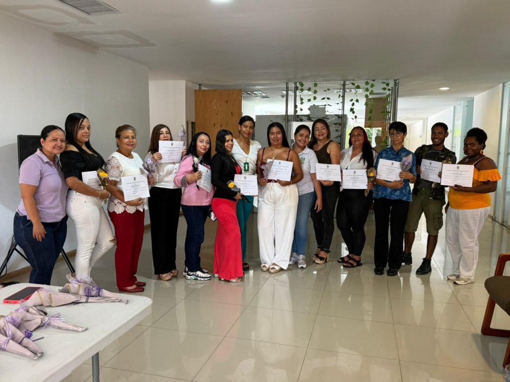 En la fotografía un grupo diverso de mujeres cuidadoras posa sonriente en un pasillo institucional sosteniendo diplomas de certificación. A la derecha, les acompaña un hombre joven. Visten ropa casual y sostienen pequeños ramos de flores, reflejando diversidad de edades y etnicidades en un evento de la Alcaldía de Palmira.