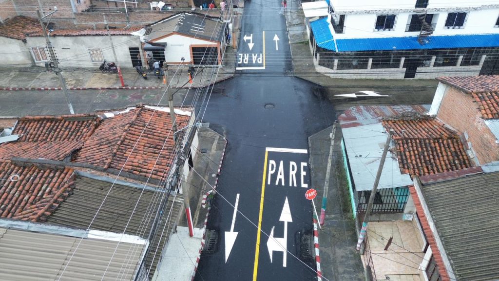 Vista aérea de una intersección vial urbana recién pavimentada y señalizada en Palmira. La calle principal muestra asfalto negro con una línea central amarilla, flechas blancas de dirección y la palabra "PARE" pintada en letras grandes y blancas sobre la calzada junto a un borde pintado de rojo y blanco.