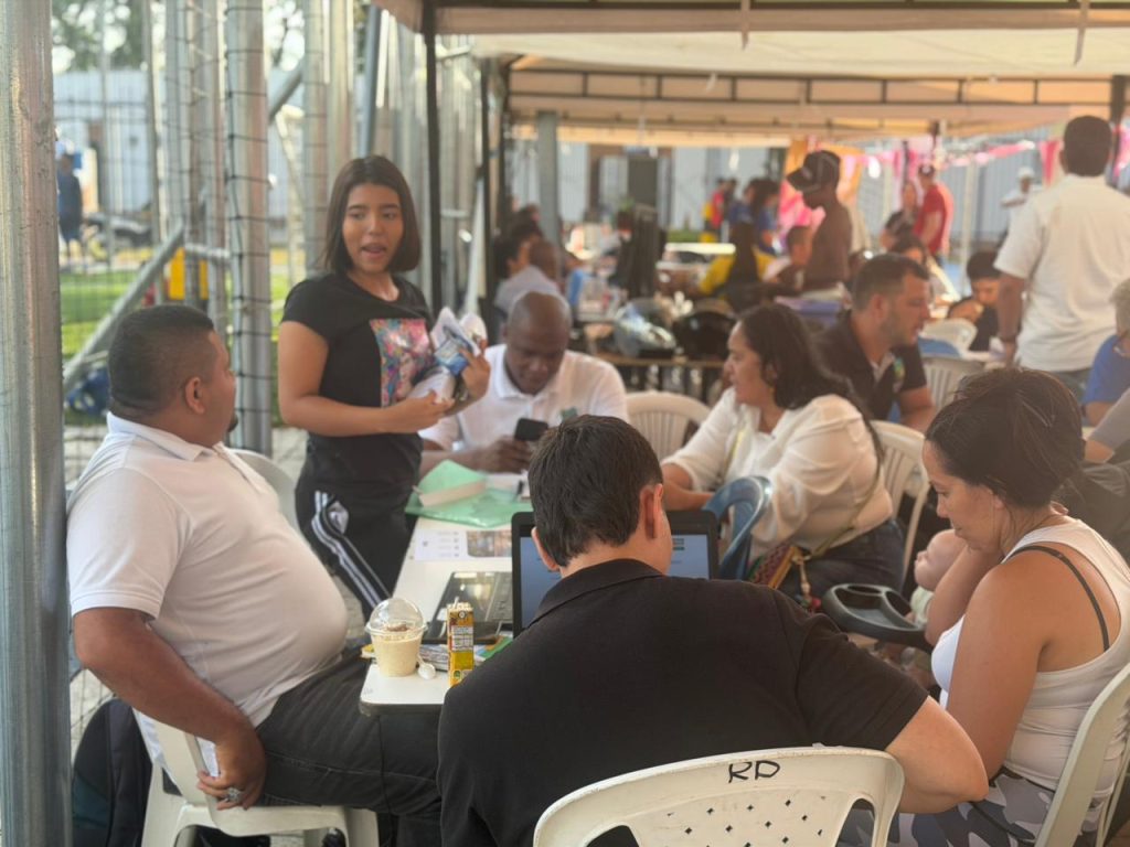En la imagen grupo diverso de personas reunido en mesas de atención bajo una carpa abierta, participando en una jornada de asesoría laboral o comunitaria con computadores y documentos