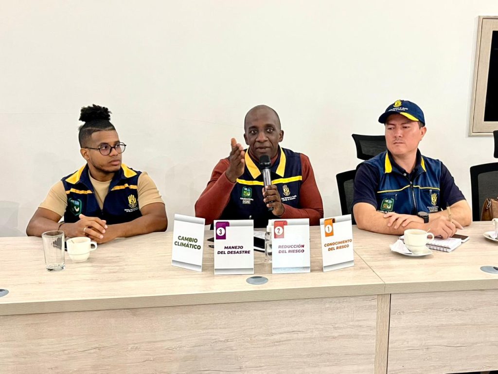 En la fotografía tres funcionarios del equipo de Gestión del Riesgo de Palmira sentados a una mesa de conferencias durante una presentación institucional. El expositor central habla por micrófono frente a cuatro carteles informativos sobre cambio climático y gestión del riesgo.