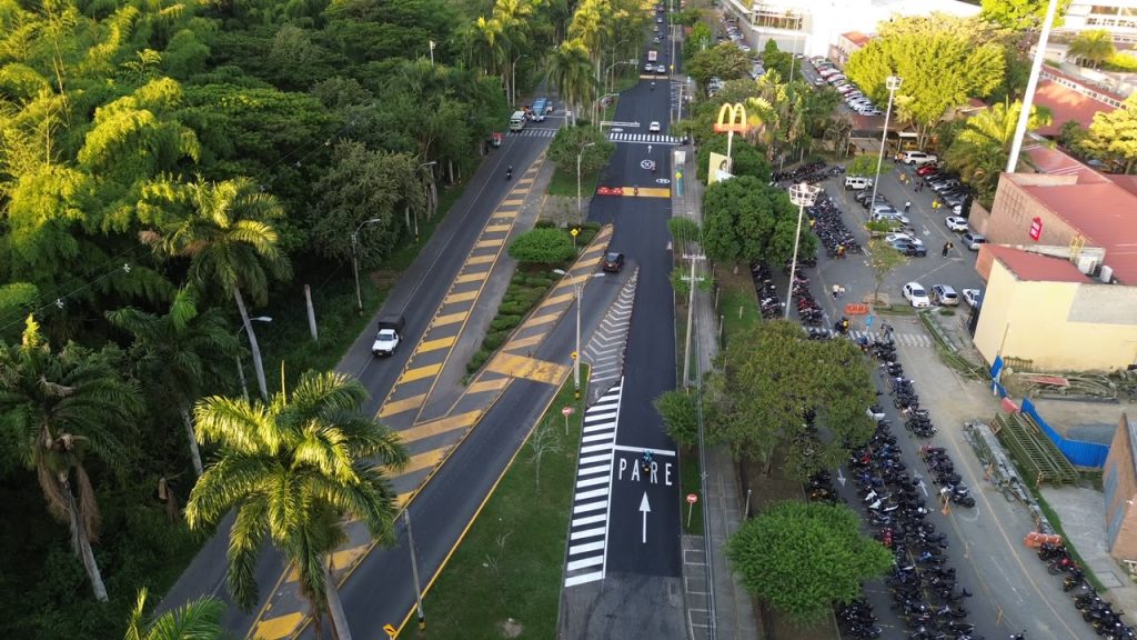 En la fotografia vista aérea de la calle 31 en Palmira, muestra una vía pavimentada con carril exclusivo de ciclorruta y señalización de "PARE". A la izquierda, un denso bosque de guaduas y palmeras; a la derecha, zonas de estacionamiento de motocicletas y locales comerciales