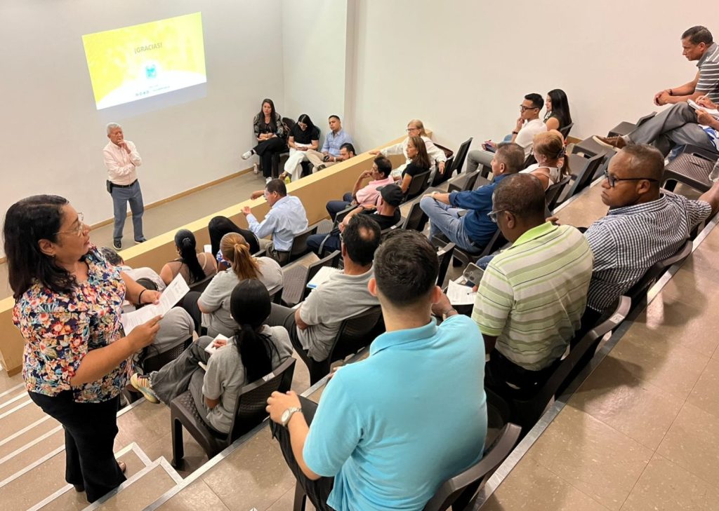 Fotografía en plano picado de una reunión o capacitación en un auditorio con gradas, donde un expositor habla frente a una pantalla proyectada y el público escucha.